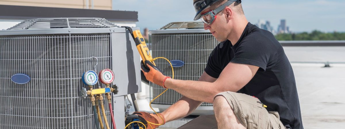 HVAC technician wearing safety gear checking rooftop air conditioning units with diagnostic gauges and a digital meter.