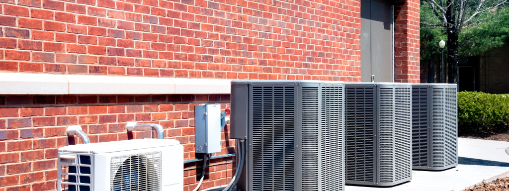 Outdoor air conditioning units installed along a red brick wall of a commercial building, illustrating HVAC solutions for efficient cooling in industrial or office settings. The image highlights energy efficiency and modern climate control systems.