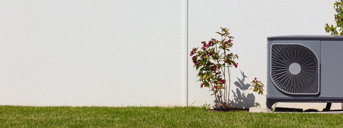 Outdoor air conditioning unit installed beside a white exterior wall with grass and a flowering shrub nearby.
