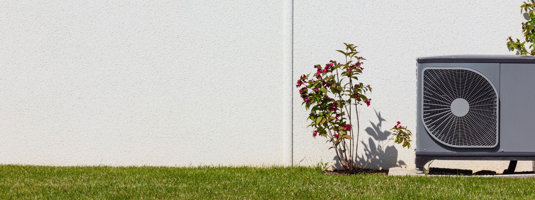 Outdoor air conditioning unit installed beside a white exterior wall with grass and a flowering shrub nearby.