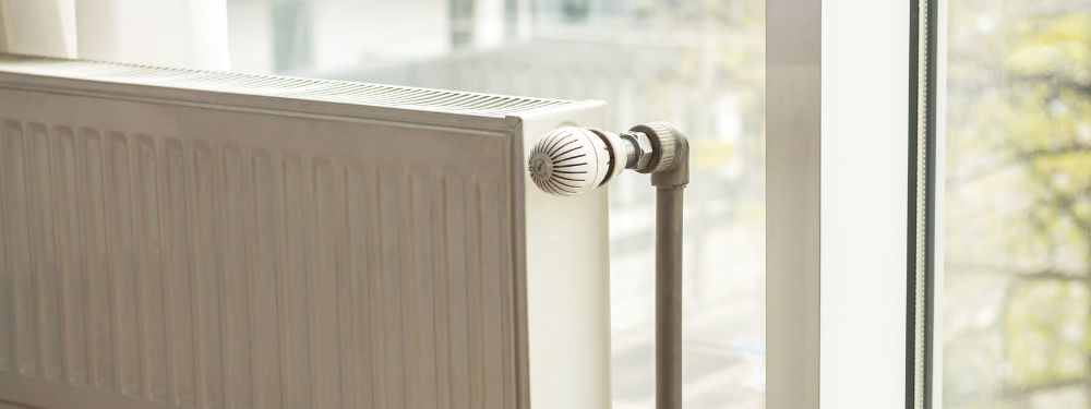 Close-up of a modern white radiator heater with a thermostat valve, positioned near a large window with a view outside.
