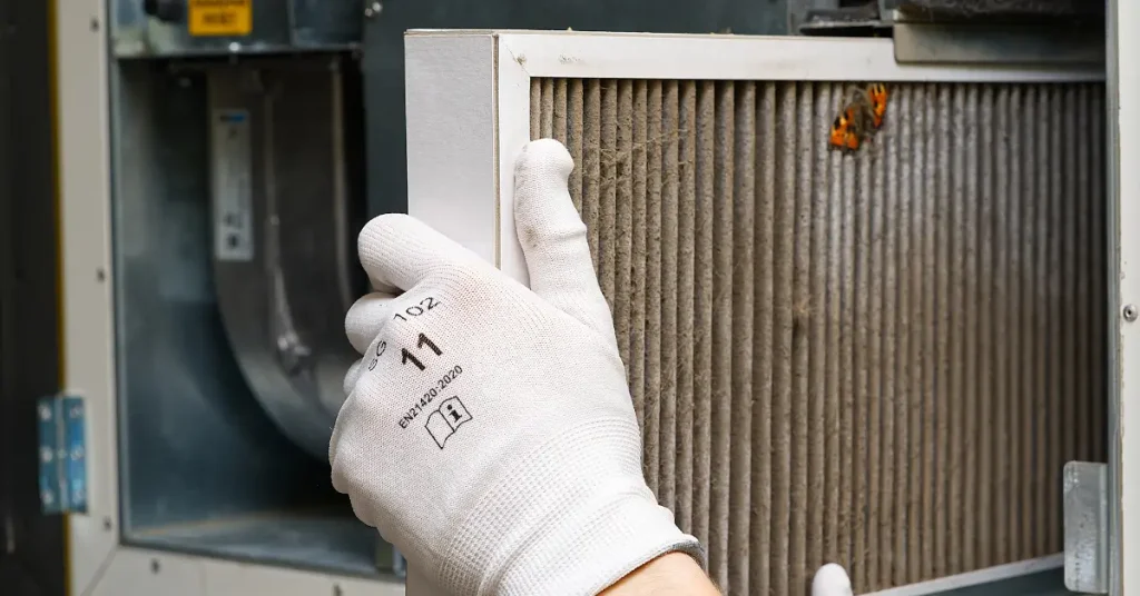 A person wearing white work gloves removes a dirty air filter from an HVAC unit, with dust buildup and a small butterfly resting on the filter.