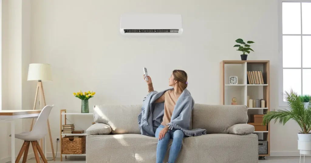A woman sitting on a sofa wrapped in a blanket points a remote control at a wall-mounted air conditioner in a bright, modern living room.