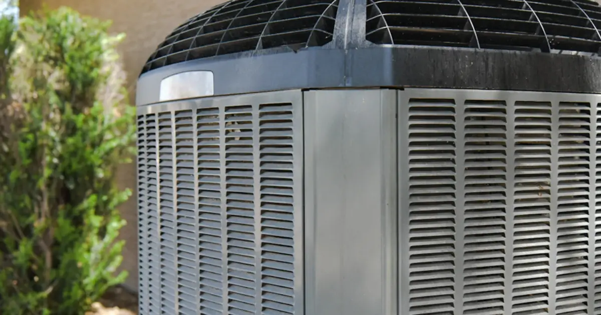 An outdoor central air conditioning unit with a metal housing and ventilation grilles, situated next to a building with greenery in the background.
