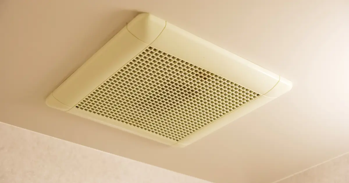 A close-up view of a beige ceiling air vent or exhaust fan with a square grid design, mounted in an indoor space.