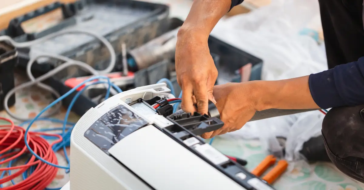 A technician’s hands working on the wiring of an indoor air conditioning unit, with tools, cables, and equipment scattered in the background.