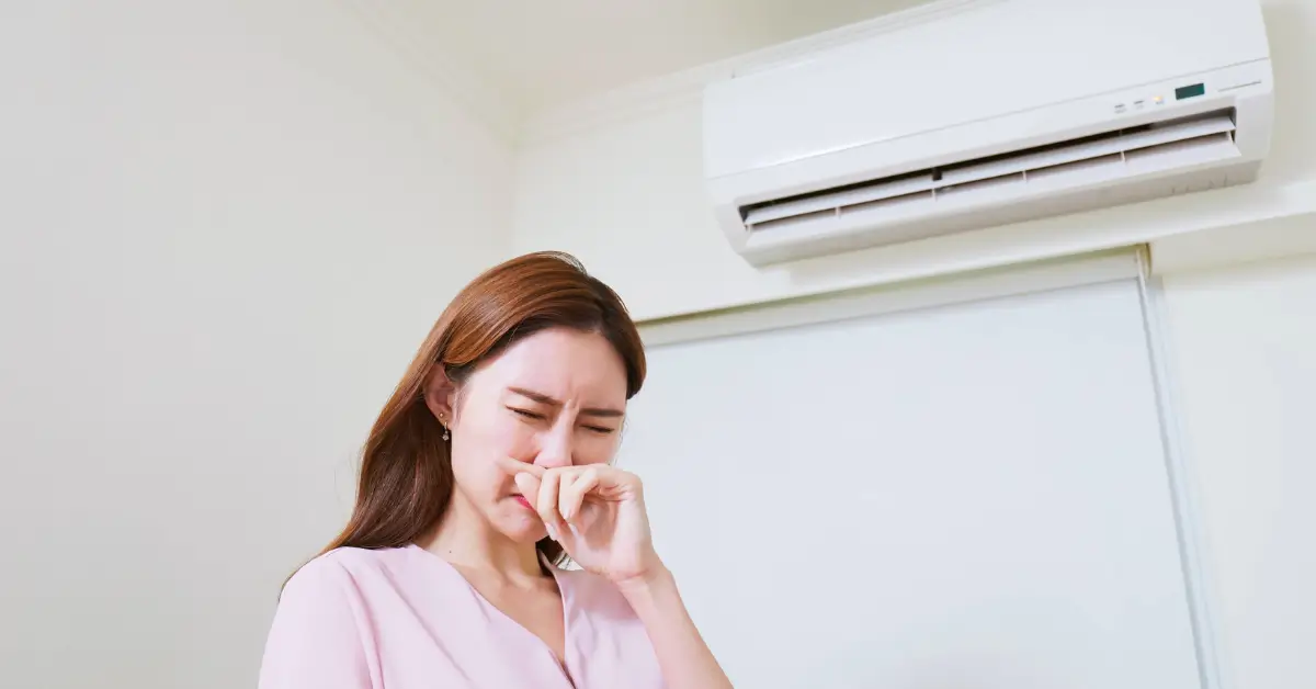 A woman covering her nose with a disgusted expression, standing beneath a wall-mounted air conditioning unit, suggesting an unpleasant odor coming from the system.