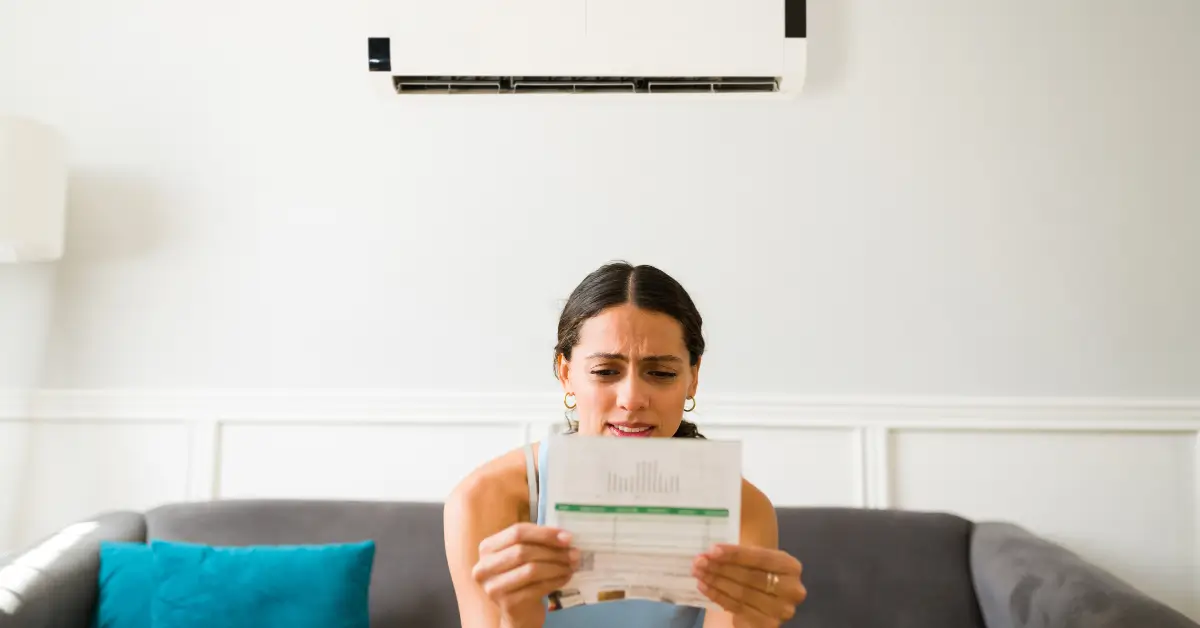A woman sitting on a couch under a wall-mounted air conditioner, looking concerned while holding and examining a utility bill.