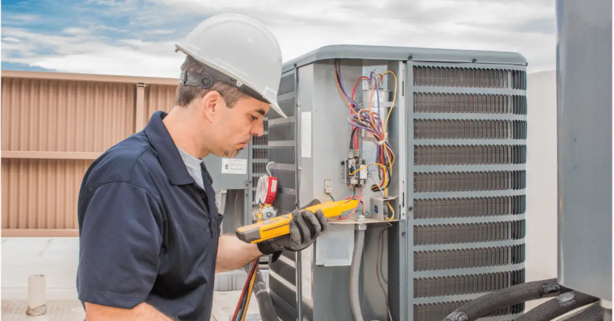 An HVAC technician wearing a hard hat and gloves using a digital multimeter to test the wiring inside an outdoor air conditioning unit with its panel open.
