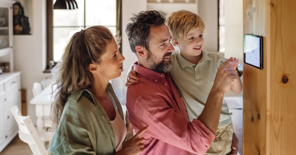 A smiling family, with a father holding his young son and the mother standing beside them, adjusting settings on a wall-mounted smart thermostat in their home.