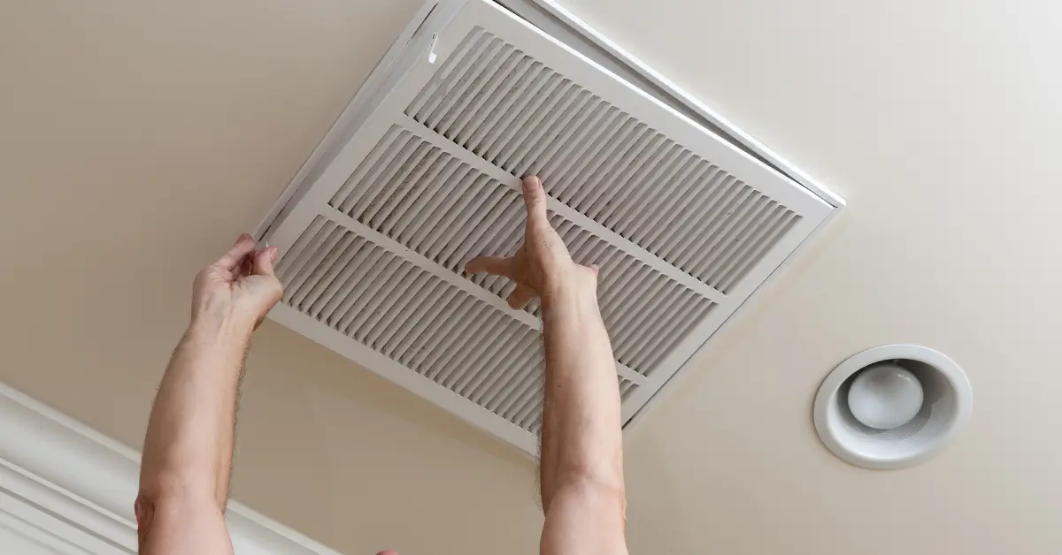 A person reaches up to open a ceiling air vent cover, exposing the HVAC air filter inside, with a recessed light fixture nearby.
