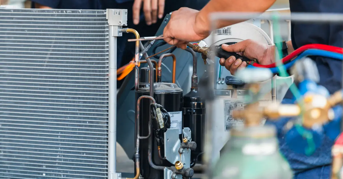 Close-up of an HVAC technician’s hands using a torch to solder copper pipes on an air conditioning unit, with visible coils, compressor components, and connected service hoses.