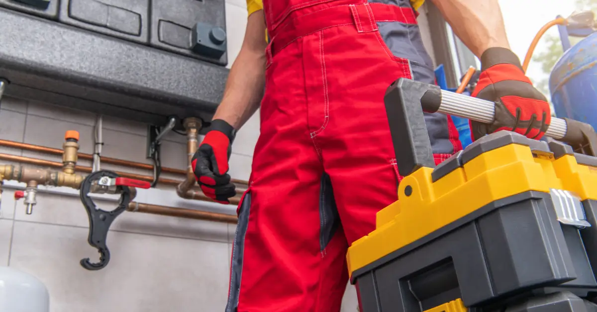 A worker in red overalls and red-and-black gloves carries a yellow and black toolbox, standing in front of plumbing and HVAC equipment with visible copper pipes and valves.