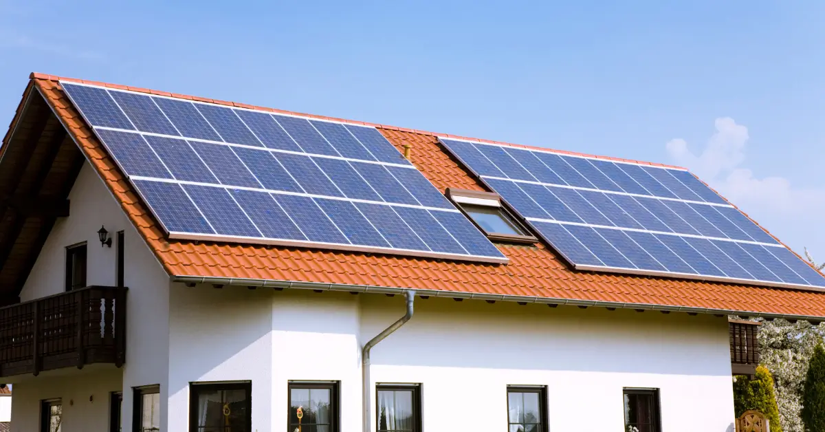 A modern house with a red tile roof is equipped with multiple large solar panel arrays, set against a clear blue sky.