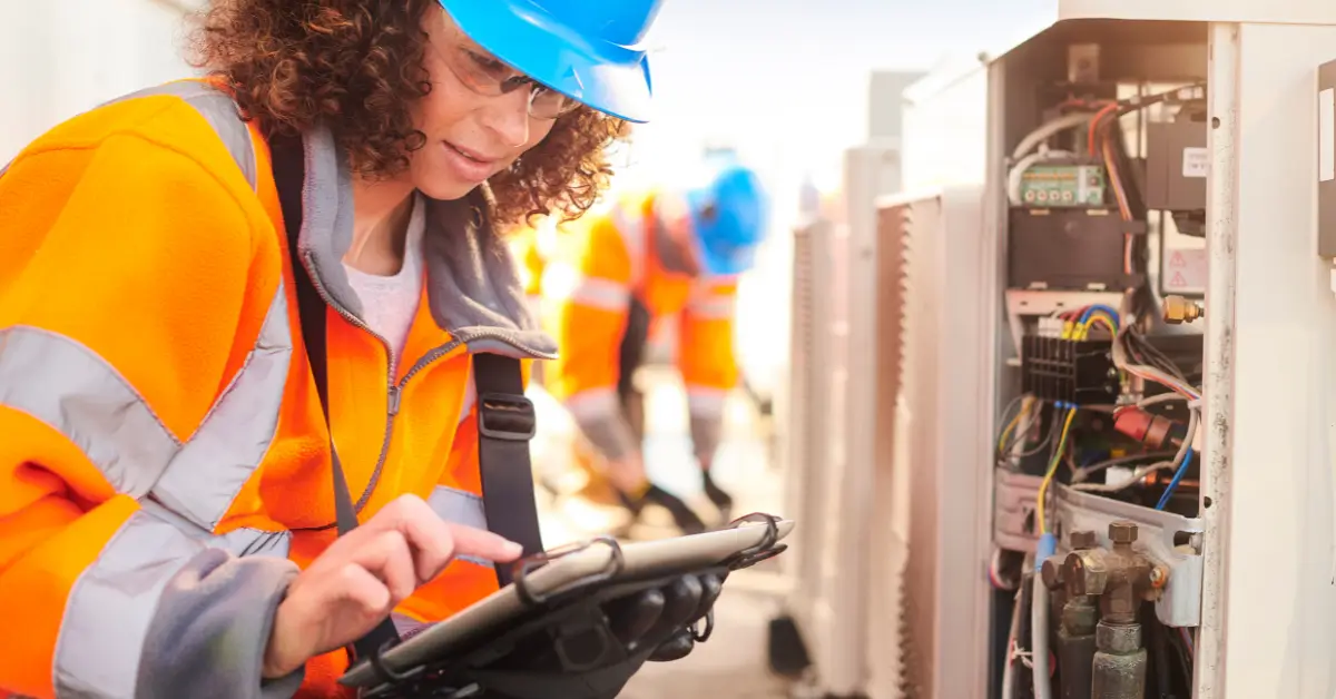 A female HVAC technician wearing a blue hard hat, safety glasses, and an orange high-visibility jacket using a tablet while inspecting an open outdoor air conditioning unit with exposed wiring.