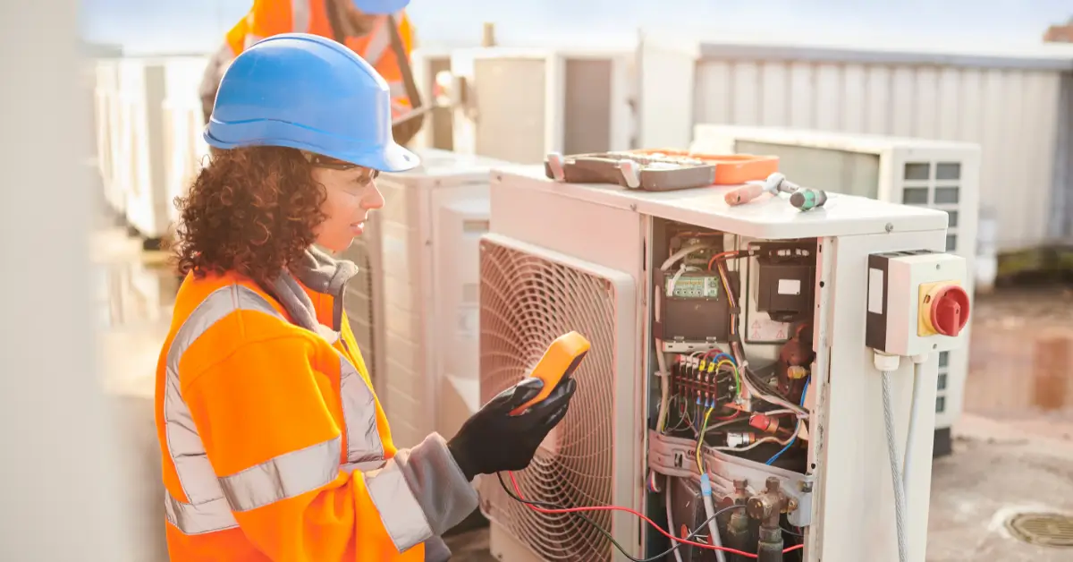 A female HVAC technician wearing a blue hard hat and orange high-visibility jacket using a handheld meter to test the wiring inside an open outdoor air conditioning unit on a rooftop.