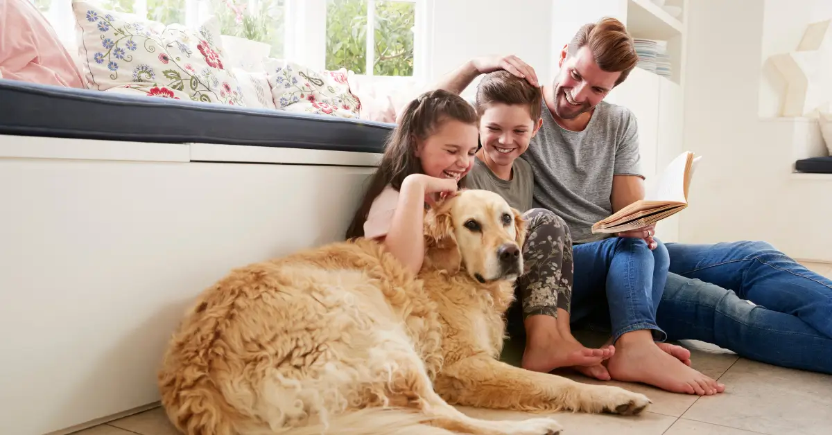 A man and two children sit on the floor smiling and laughing together while petting a relaxed golden retriever. The man is holding an open book, and they are sitting next to a cushioned window seat with floral pillows in a bright, cozy room.