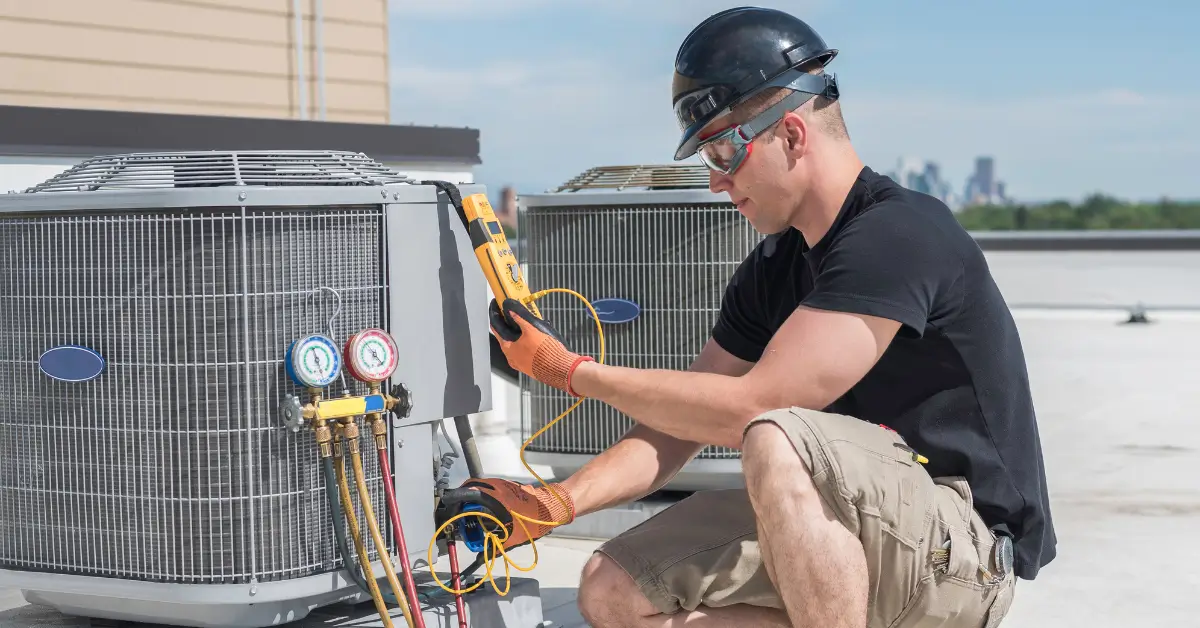 An HVAC technician wearing a hard hat, safety glasses, and gloves works on an outdoor air conditioning unit, using electrical testing tools and gauges to check the system’s performance. Several other similar units are visible in the background on a rooftop.