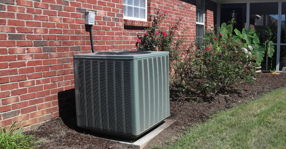 An outdoor HVAC unit sits on a concrete pad next to a red brick house, with electrical wiring connected above it. Flowering bushes and green plants grow along the side of the house, and a grassy lawn is visible in the foreground.