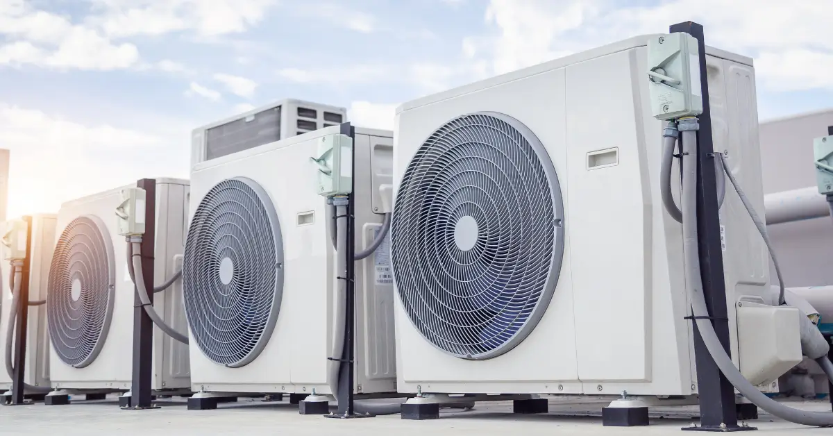 A row of large outdoor HVAC units with prominent circular fans is mounted on a rooftop, connected by electrical conduits and supported by metal stands, with a bright sky in the background.